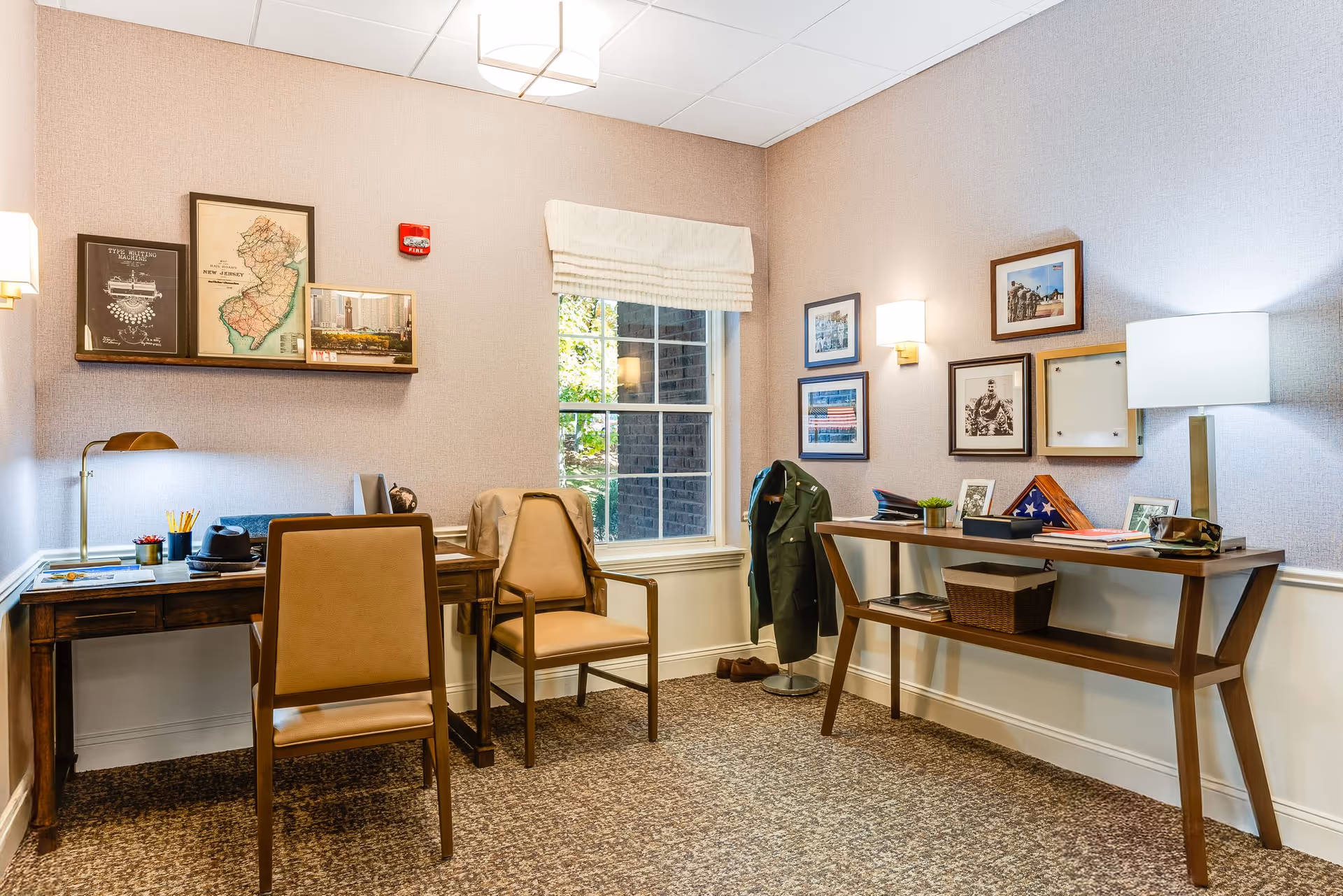 A cozy senior living office/study area with two desks, chairs, framed photos on the walls, and a window.