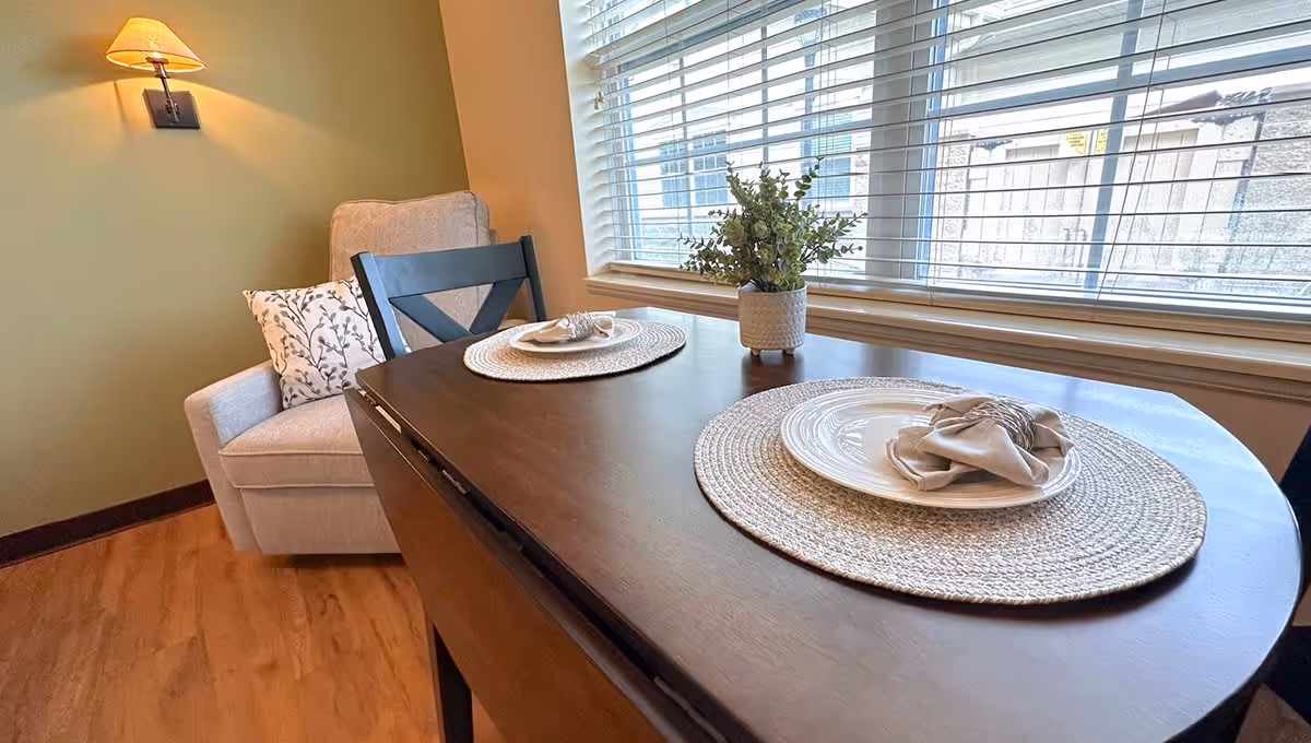 A small dining area with a dark wooden table set for two with white plates, beige napkins, and woven placemats. A potted plant is placed in the center of the table. Next to the table is a beige upholstered armchair with a decorative pillow. A wall-mounted lamp with a beige shade is on the green wall behind the chair. A large window with white blinds lets in natural light.