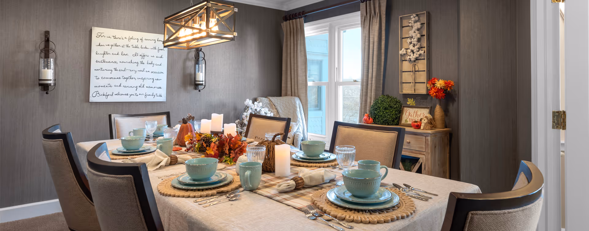 A warmly decorated dining room with a rectangular table set for six people. The table features light blue dishware, glasses, silverware, and autumn-themed centerpiece decorations including candles and small pumpkins. The room has gray walls, a window with beige curtains, a wooden sideboard with decorative items, and a modern rectangular chandelier hanging above the table.