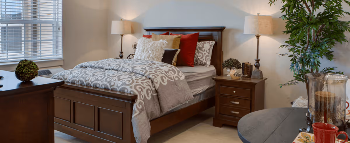 A cozy bedroom with a wooden bed frame featuring a patterned gray and white comforter, multiple pillows in red, white, and brown, a wooden nightstand with a lamp and decorative items, a window with blinds, a wooden dresser with a small green plant, and a round table with candles and a red cup, along with a tall green plant in the corner.
