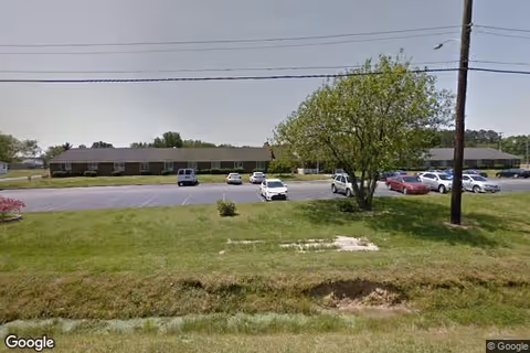Single-story retirement building front with a parking lot, several cars, a tree and grassy foreground under power lines.