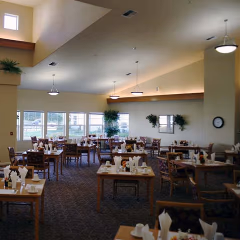 A spacious dining room with multiple wooden tables and chairs arranged neatly. Each table is set with white napkins, cups, and small flower arrangements. Large windows allow natural light to fill the room, and there are plants and wall decorations on the far wall. The ceiling has recessed lighting and hanging light fixtures.
