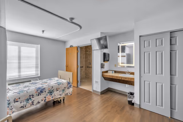 A bright and clean nursing home bedroom with a single bed covered in a floral bedspread, a large window with blinds, a wall-mounted TV, a sink with a mirror and soap dispenser, a trash bin, and an open door leading to a tiled bathroom with a shower.