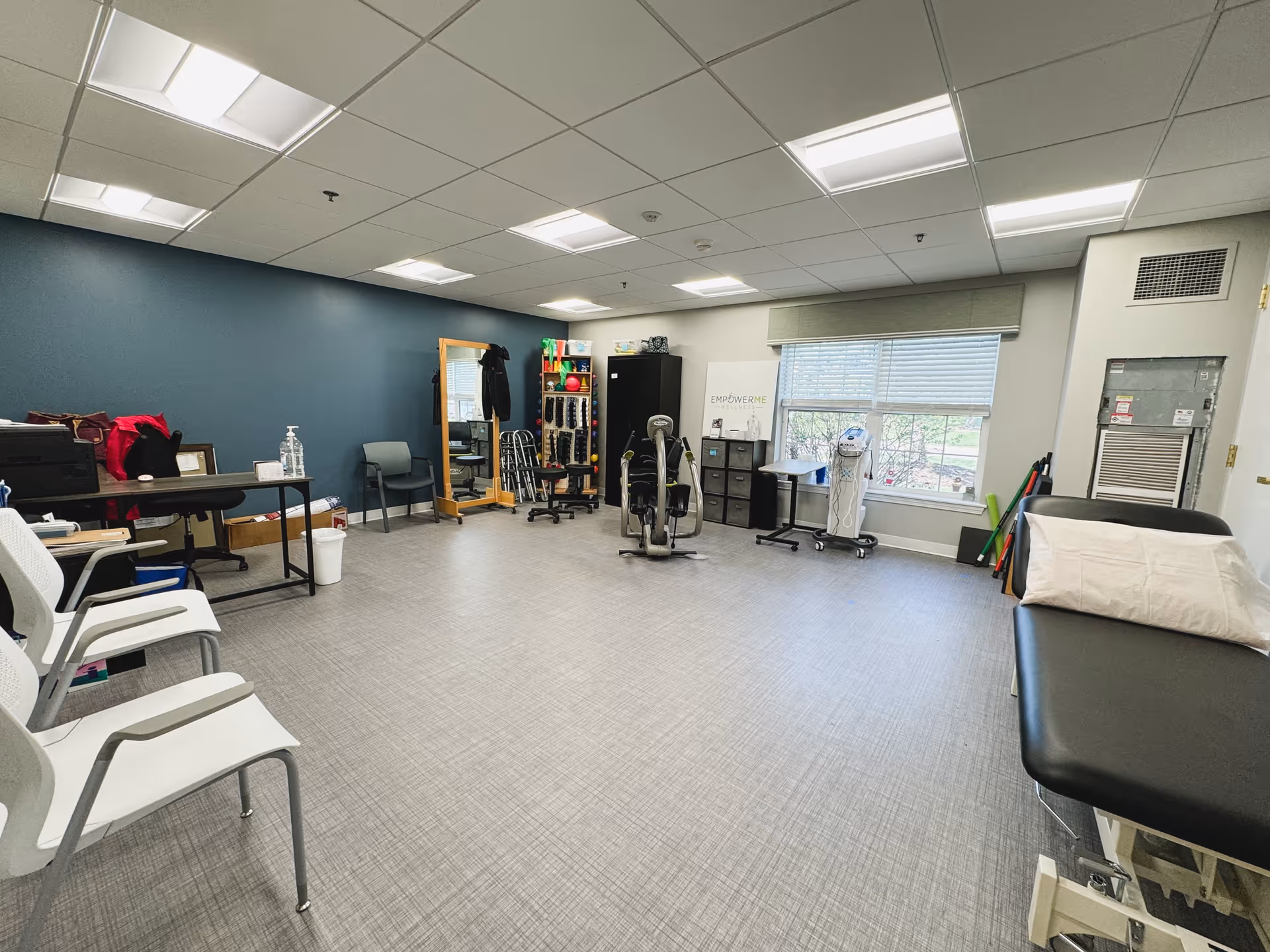 A spacious therapy or rehabilitation room with exercise equipment, chairs, a treatment table with a pillow, a large mirror, and a window letting in natural light. The room has a blue accent wall, gray flooring, and overhead fluorescent lighting.