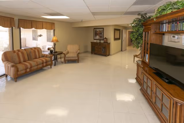 A spacious living room area in Mountain View Care Center featuring a striped sofa and an armchair near large windows with valance curtains. There is a wooden side table with a lamp and flowers, a wooden cabinet with framed artwork above it, and a large wooden entertainment center with a flat-screen TV and books. The floor is tiled, and there is a hallway leading to other rooms.