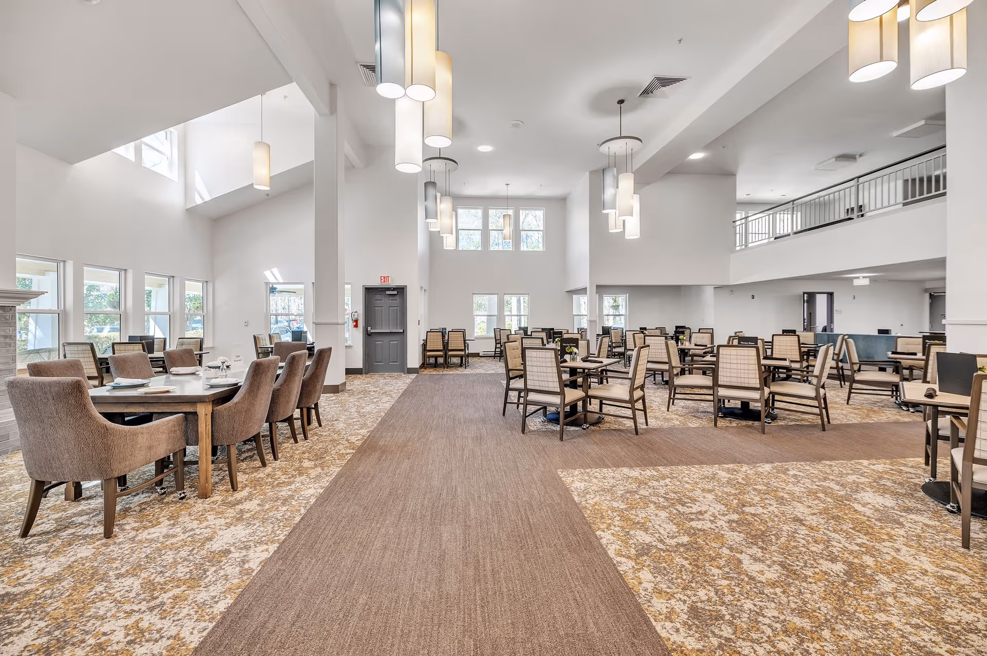 Large bright senior living dining and common area with multiple tables and chairs, high ceilings, and pendant lighting.