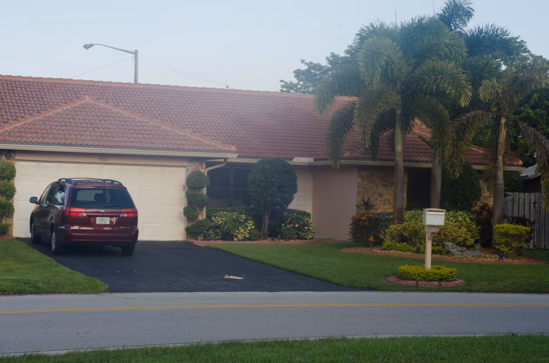 Single-story house with a red tiled roof, a driveway with a red minivan parked, well-maintained green lawn, trimmed bushes, palm trees, and a white mailbox in front.