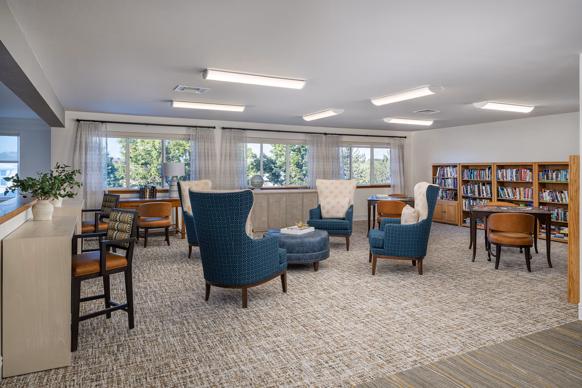 Well-lit communal lounge with upholstered armchairs and an ottoman arranged around a low table, large windows and bookshelves in the background.