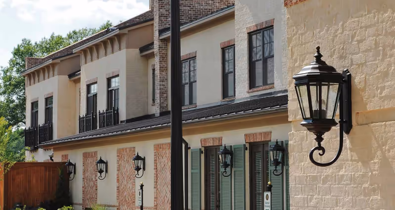 Exterior view of a senior living facility building with beige and brick walls, black lantern-style wall lights, green shutters on windows, and a wooden gate. Trees and greenery are visible in the background.
