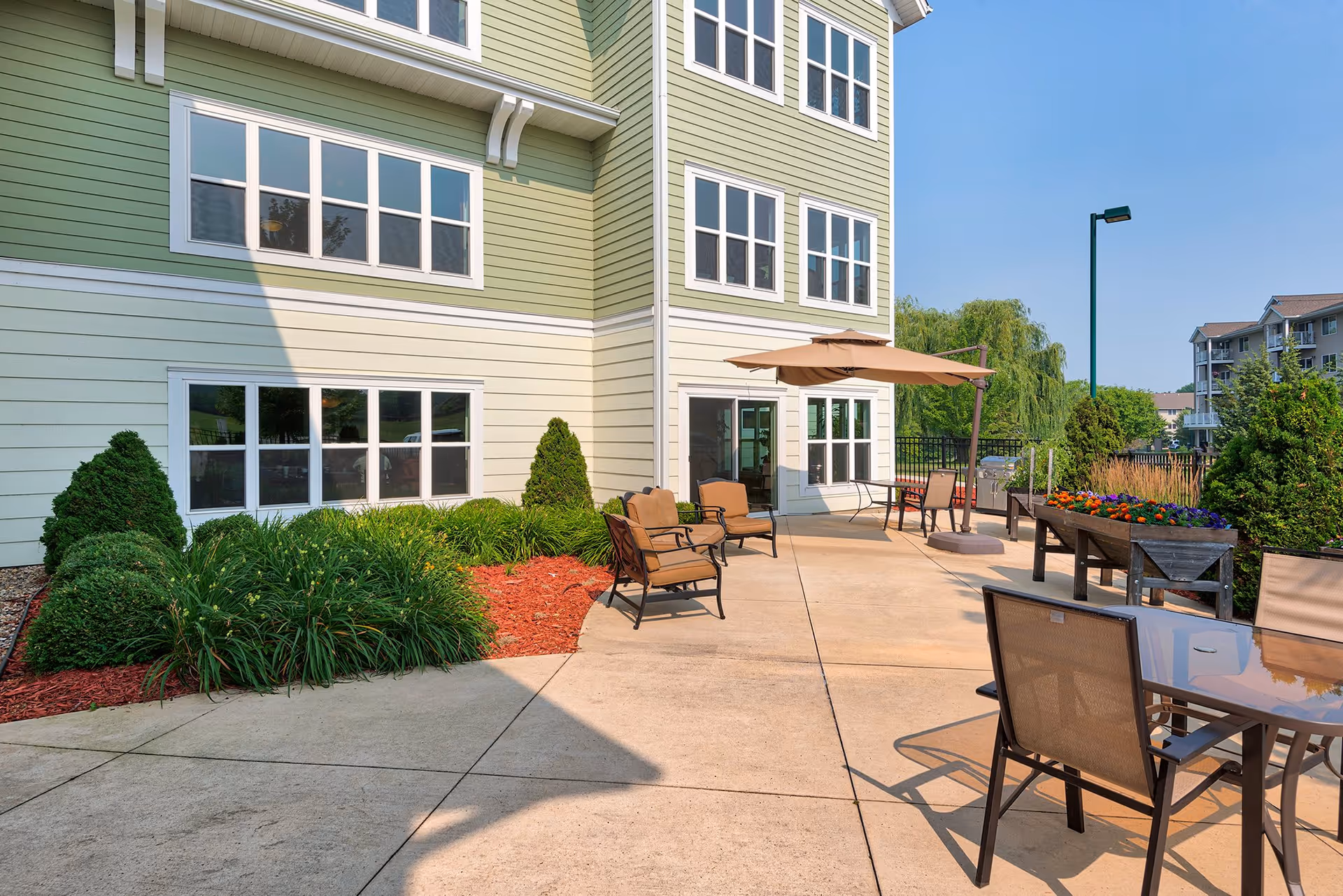 Outdoor patio area at a senior living facility with several cushioned chairs, a table with chairs, an umbrella, flower planters, and well-maintained landscaping next to a green and beige building under a clear blue sky.