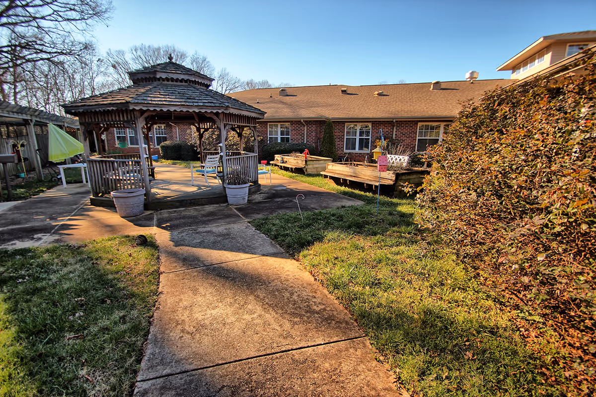 Outdoor area of a senior living facility featuring a wooden gazebo with chairs inside, surrounded by a concrete pathway, grass, bushes, and raised garden beds. The background shows a brick building under a clear blue sky.