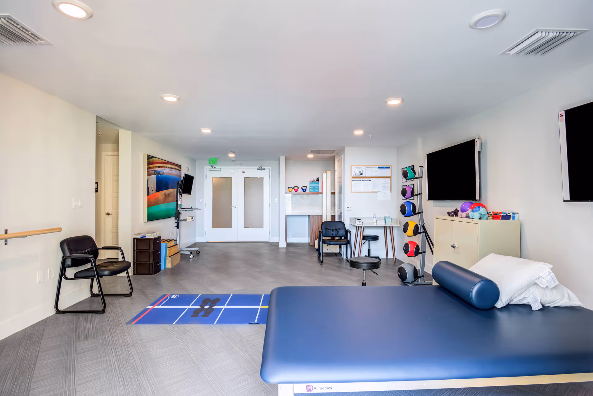 A bright and spacious physical therapy or rehabilitation room with a padded treatment table in the foreground, exercise balls on a rack, two wall-mounted televisions, chairs, and a colorful shuffleboard game mat on the floor. The room has white walls, recessed lighting, and a double door at the far end.