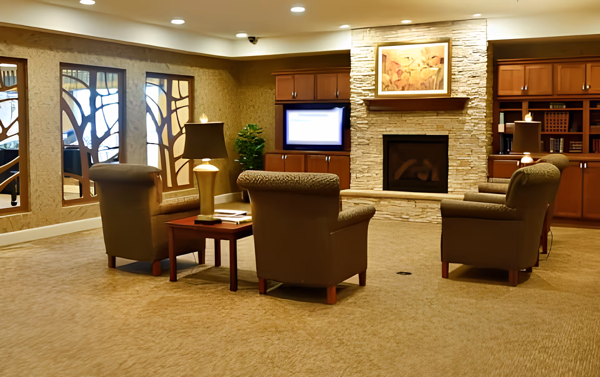 A cozy living room area with four cushioned armchairs arranged around a wooden coffee table with a lamp and books. The room features a stone fireplace with a framed painting above it, built-in wooden cabinets, and decorative wall mirrors with artistic wooden frames. The carpeted floor and warm lighting create a comfortable atmosphere.