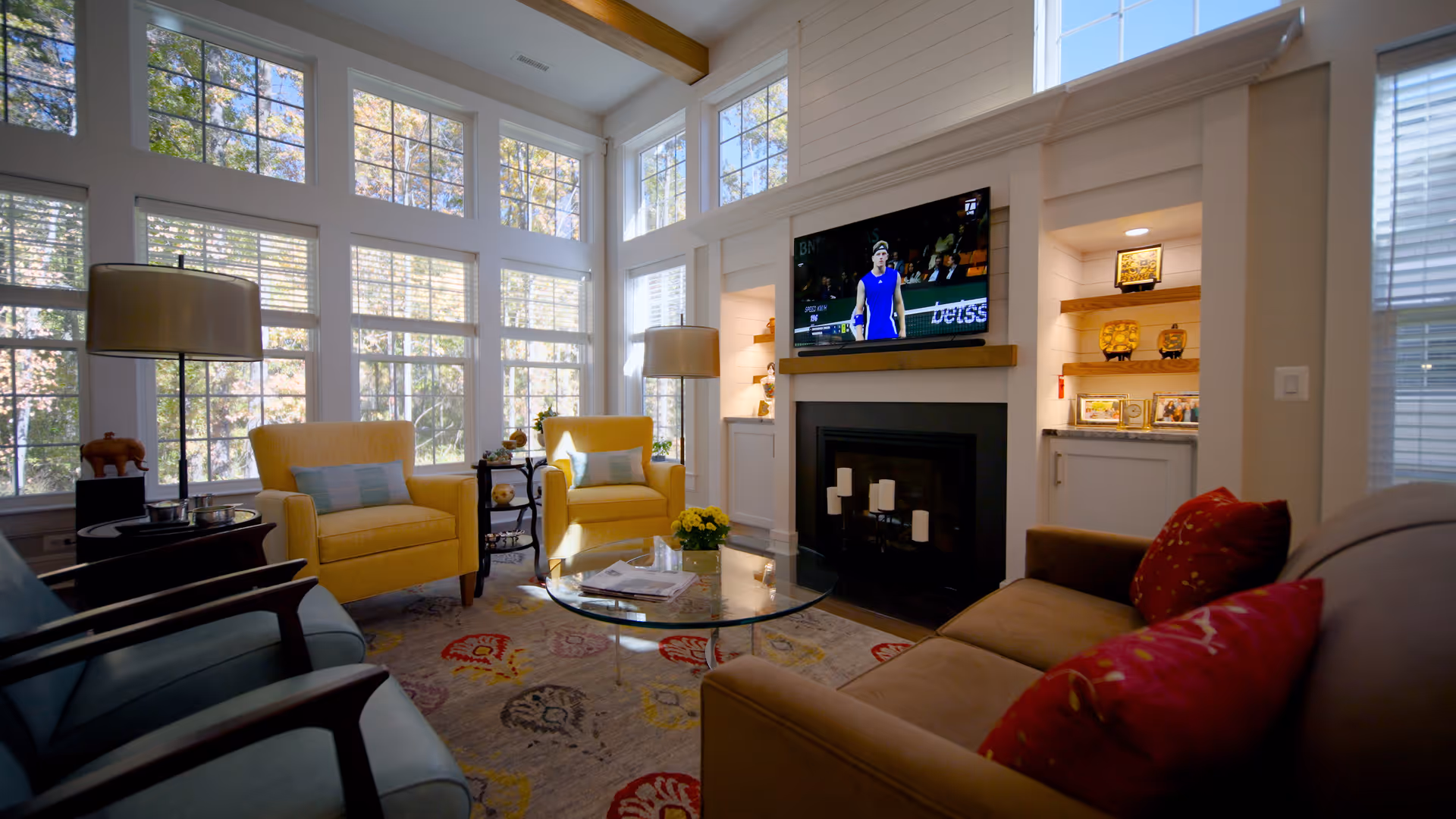 Sunlit living room with tall windows, yellow armchairs, a fireplace with a mounted TV, and sofas around a glass coffee table.