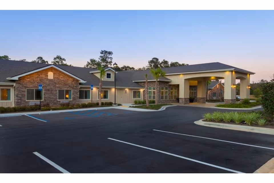 Front exterior of a single-story senior living facility with a covered porte-cochère, palm trees, and an empty parking lot at dusk.