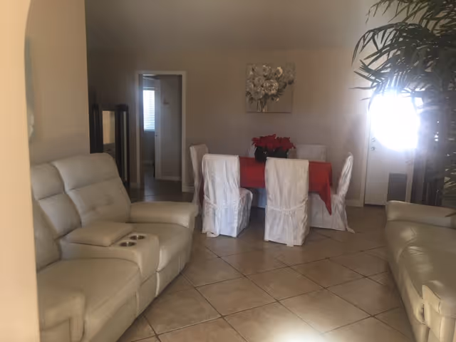 Interior view of a living and dining area with beige tiled floors. There are two cream-colored recliner sofas on either side of the room. In the center, there is a dining table covered with a red tablecloth and surrounded by six white slipcovered chairs. A floral painting hangs on the wall above the dining table. A potted plant is partially visible on the right side near a door with bright sunlight coming through.