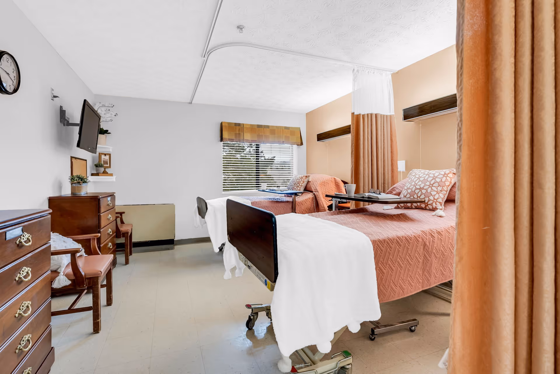 A shared patient room in a healthcare facility with two hospital beds, each covered with a pink quilted blanket and a white towel at the foot. The beds have adjustable overbed tables with items on them. There is a window with blinds and a valance, a wall-mounted TV, wooden dressers, chairs, and a privacy curtain partially drawn between the beds.