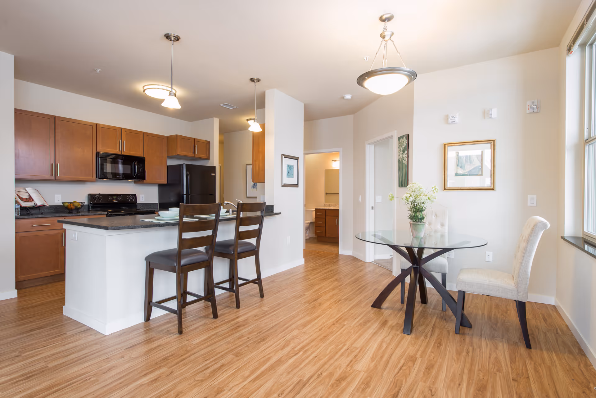 A bright and modern kitchen and dining area with wooden floors. The kitchen features wooden cabinets, a black refrigerator, microwave, and stove. There is a white island with two dark wooden chairs. The dining area has a round glass table with two upholstered chairs and a vase with flowers. The walls are light-colored with framed artwork and a window letting in natural light.