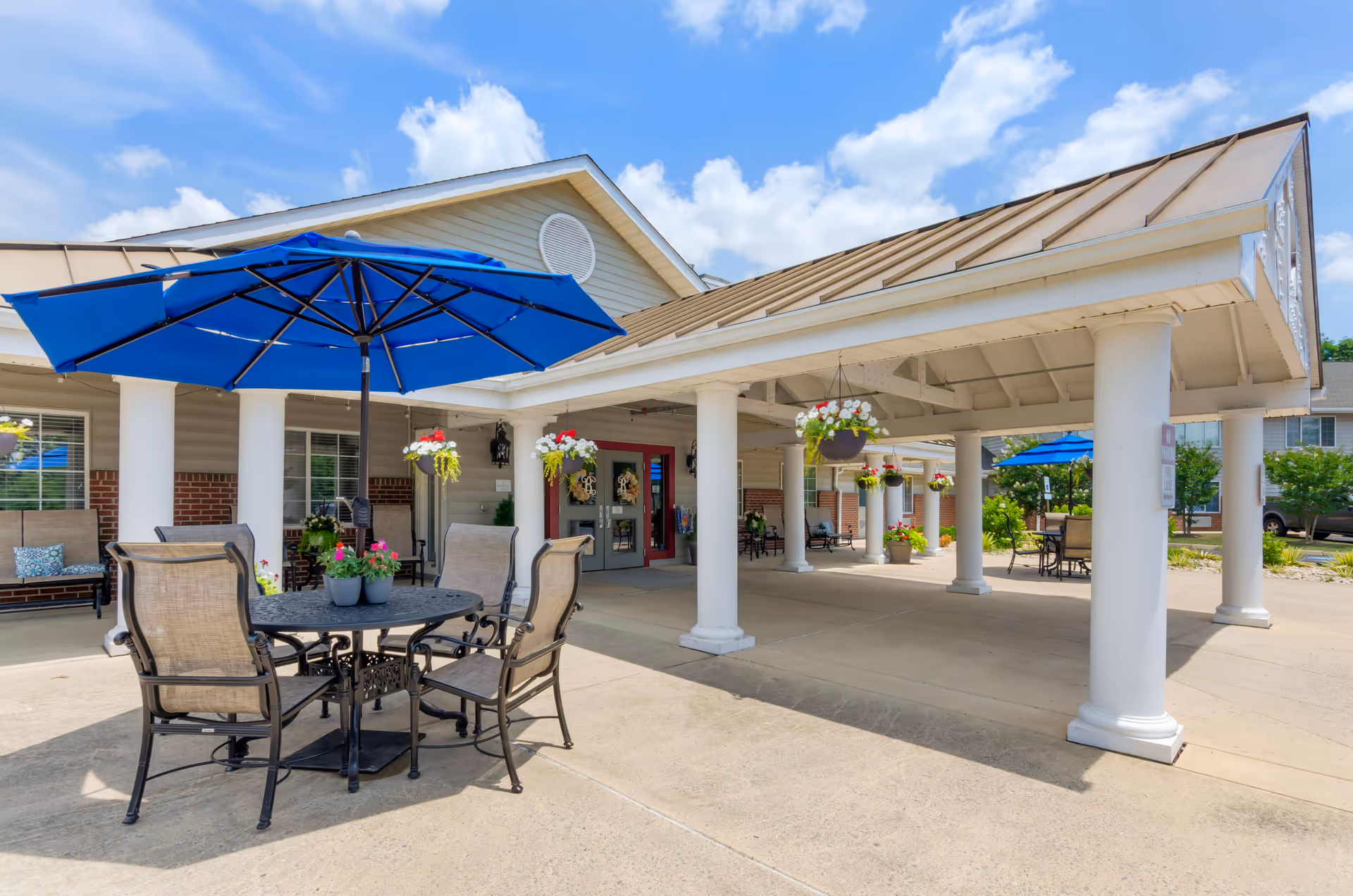 Outdoor seating area at Brookdale Staunton with a round metal table, four chairs, and a large blue umbrella. The area is shaded by a covered porch supported by white columns, decorated with hanging flower baskets. The building exterior features beige siding, brick accents, and a red entrance door under a partly cloudy sky.