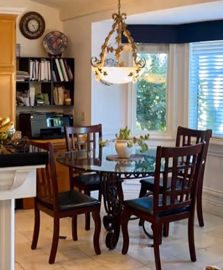 A cozy dining area with a round glass table surrounded by four wooden chairs with black cushions. Above the table hangs an ornate chandelier with floral designs. In the background, there is a window with blinds partially open, letting in natural light. To the left, there is a kitchen counter and a shelving unit with books, a globe, and a printer.