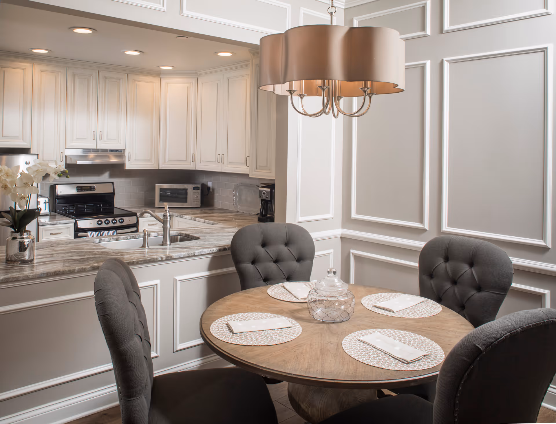 A cozy dining area with a round wooden table set with four placemats and napkins, surrounded by four tufted gray chairs. Behind the dining area is a kitchen with white cabinetry, a stainless steel stove, microwave, coffee maker, and a marble countertop with a sink. A modern chandelier hangs above the table, and the walls have decorative molding.