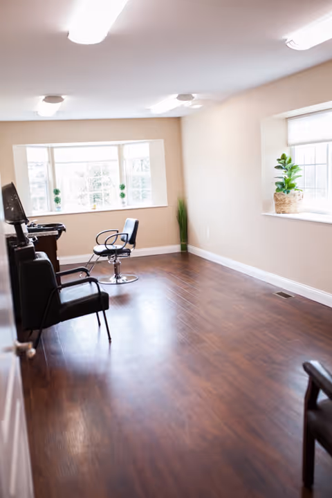 A bright room with wooden flooring and beige walls featuring two black salon chairs and a styling station near a large window with natural light. There are small green plants on the window sill and a tall plant in the corner.