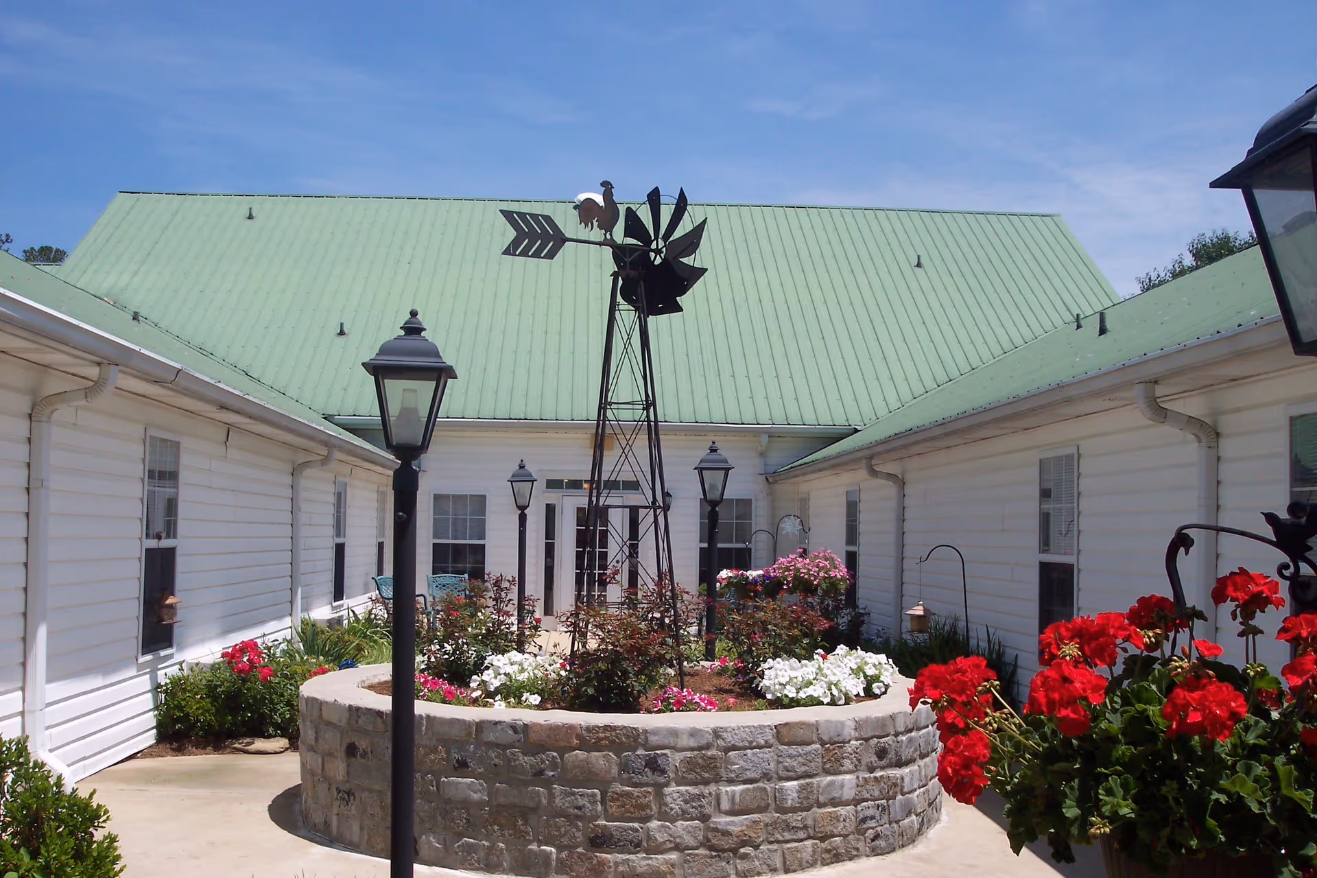 Outdoor courtyard area of Kimberly Personal Care Home featuring a circular stone flower bed with colorful flowers and a decorative windmill with a rooster on top. The courtyard is surrounded by white buildings with green metal roofs and black lamp posts.