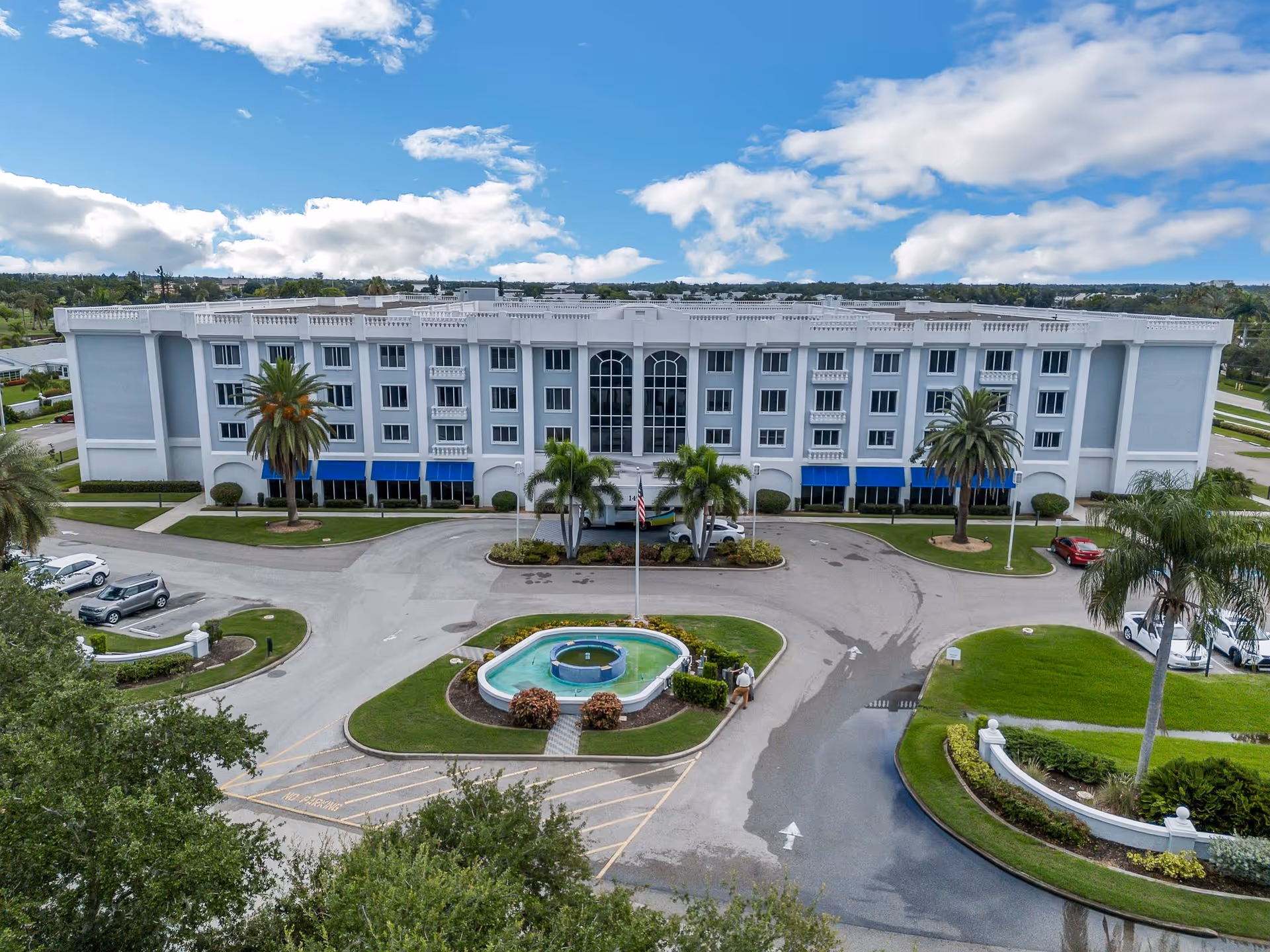 Front exterior view of a large, white multi-story building with blue awnings over the ground floor windows. The building is surrounded by a circular driveway with a small fountain and landscaped greenery in the center. Palm trees and other plants are visible around the property under a partly cloudy blue sky.