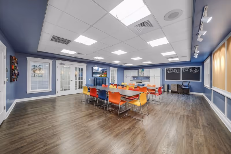 A spacious, well-lit room with blue walls and wood flooring featuring a long table surrounded by colorful chairs in red, orange, yellow, and blue. The room has a kitchenette area with white cabinets and a countertop, a wall-mounted TV, two large windows, French doors, and two blackboards with drawings on the far wall. There is also a desk with a chair and some bulletin boards on the right wall.