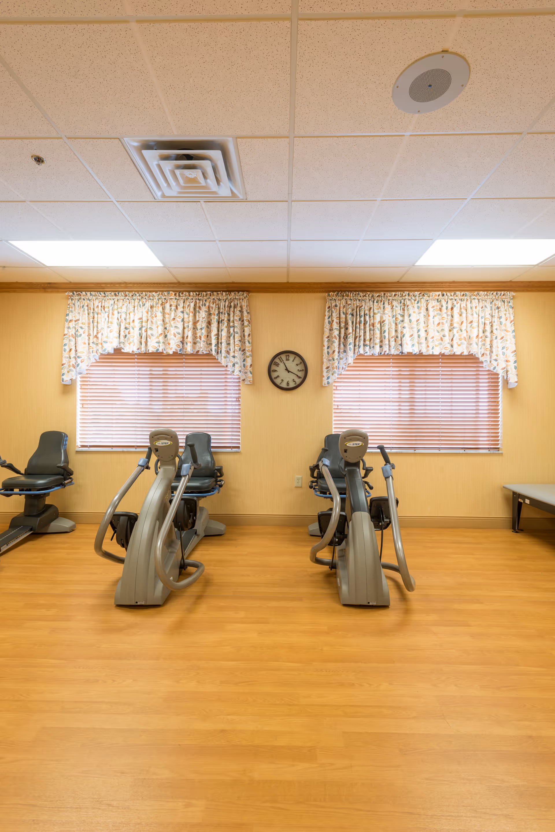 Small exercise room with recumbent stationary bikes lined up facing two curtained windows and a wall clock.