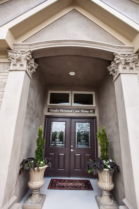 Entrance to Tender Personal Care Home #2 featuring double dark brown doors with glass panels, a doormat, two large decorative planters with green and purple plants on either side, and an arched architectural detail above the doorway.
