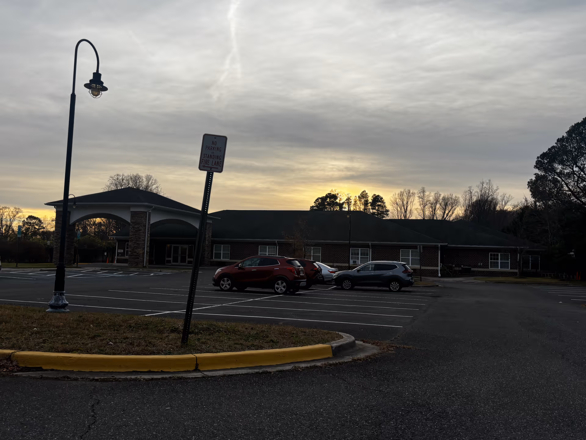 Exterior view of a single-story brick building with a covered entrance and a parking lot in front. Several cars are parked, and a streetlamp and a 'No Parking Standing Fire Lane' sign are visible. The sky is cloudy with the sun setting in the background.