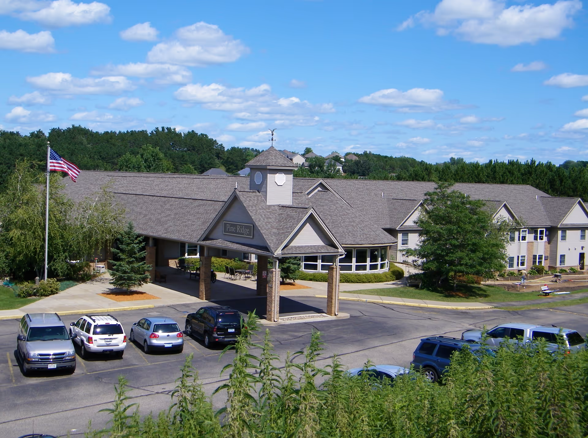 Exterior view of Pine Ridge Assisted Living facility showing a large building with a covered entrance, several parked cars, an American flag on a flagpole, and surrounding greenery under a blue sky with scattered clouds.