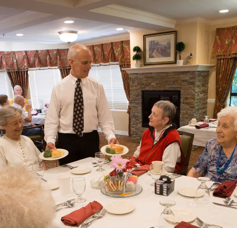 A waiter serving plates of food to elderly women seated around a dining table in a well-lit room with a stone fireplace and floral curtains.