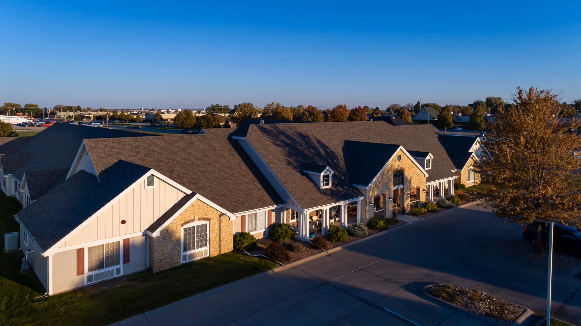 Aerial view of Homestead Assisted Living of Kearney building with a combination of stone and siding exterior, multiple windows, and a large driveway surrounded by greenery and trees under a clear blue sky.