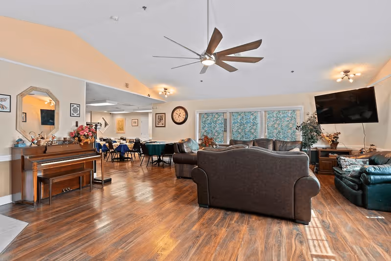 A spacious senior living facility common area with wooden flooring, a ceiling fan, and multiple seating arrangements including leather sofas and armchairs. There is a piano with decorative items on it, a wall clock, a large flat-screen TV mounted on the wall, and windows with patterned curtains. In the background, a dining area with tables and chairs is visible.