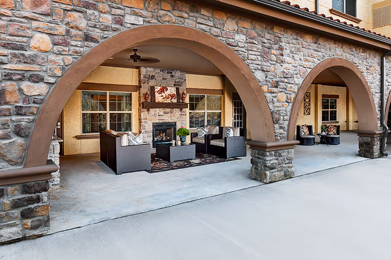 Outdoor covered patio area with stone arches and a seating arrangement including cushioned chairs and a coffee table in front of a stone fireplace, decorated with plants and artwork.