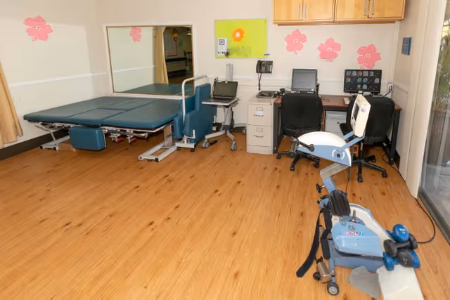 A small therapy/rehab room with a treatment table, exercise bike, desks and computers against a wall decorated with pink flower decals.