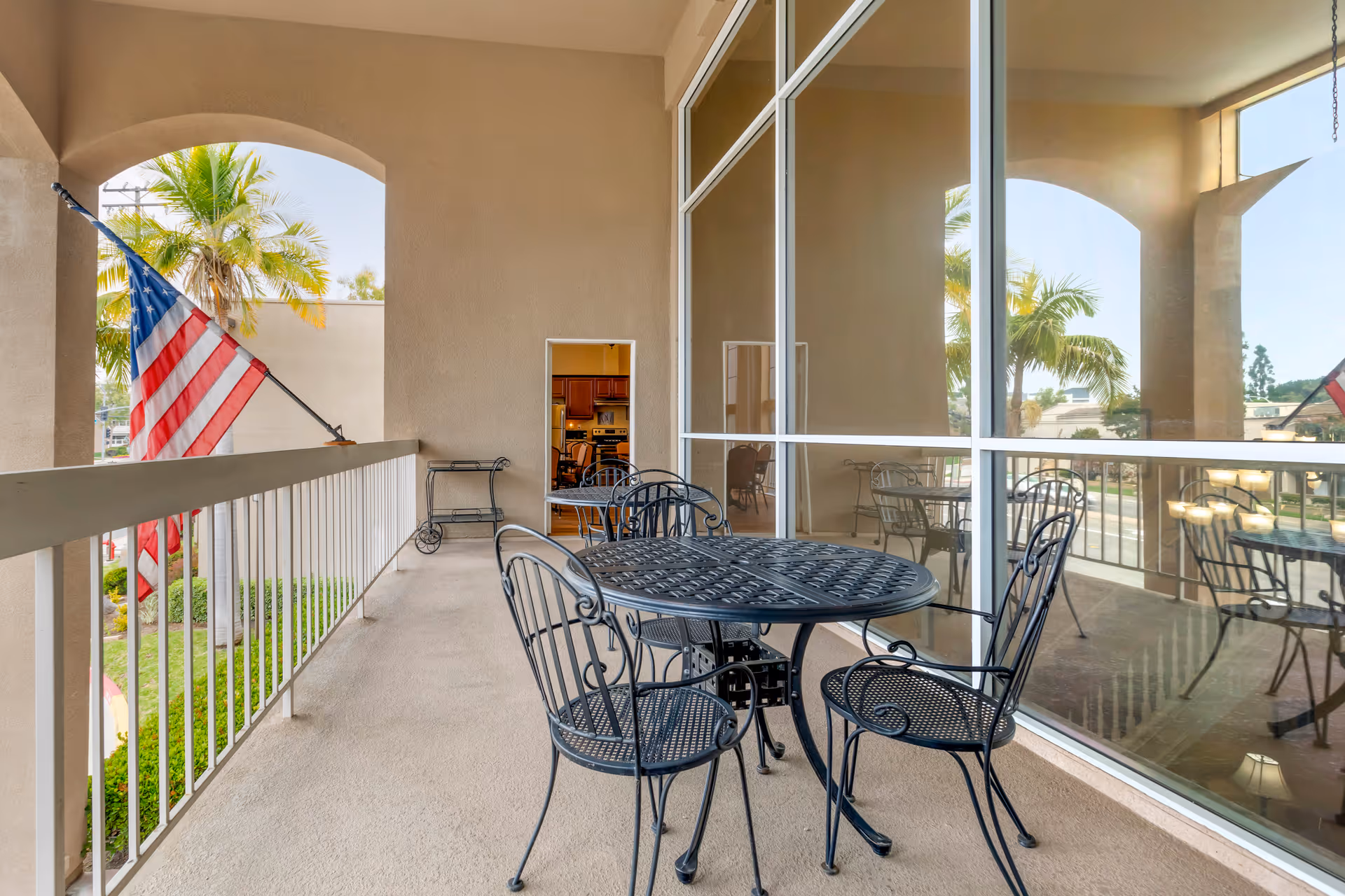 Outdoor covered balcony area with a black metal table and four matching chairs. An American flag is mounted on the railing. Large windows reflect the balcony furniture and palm trees are visible outside.