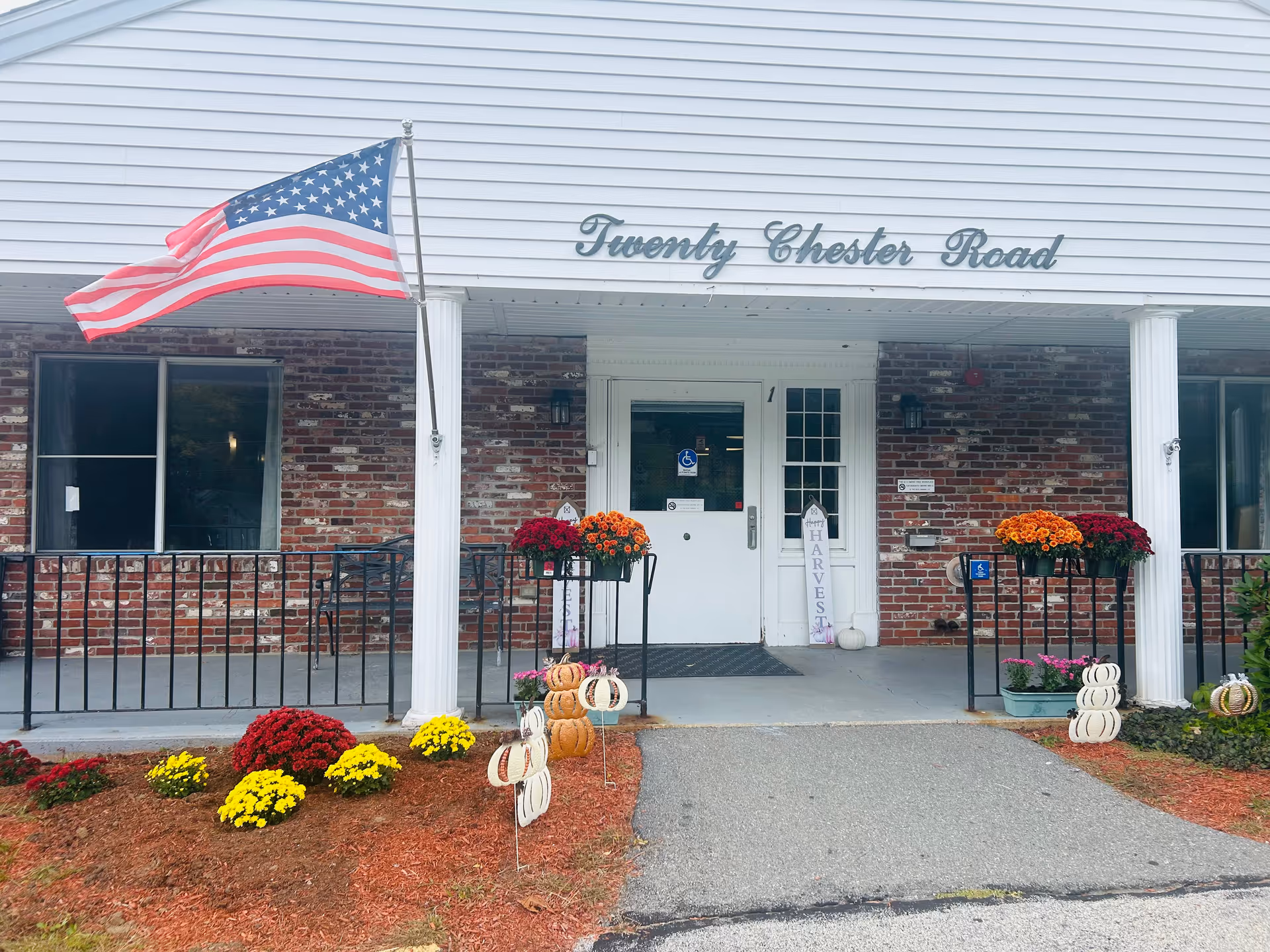 Entrance of a brick building with white pillars and a white door. Above the door is a sign that reads 'Twenty Chester Road'. There is an American flag on a pole to the left of the entrance. Flower pots with orange, red, and yellow flowers decorate the entrance area, along with small pumpkin decorations and a sign that says 'Harvest'.