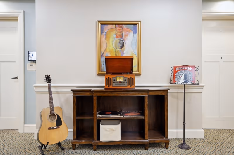 A wooden cabinet with an open vintage record player on top, a framed painting of a guitar hanging on the wall above it, a guitar standing on a stand to the left, and a music stand holding a 'Godspell' album cover to the right. The setting appears to be a hallway with two white doors on either side.