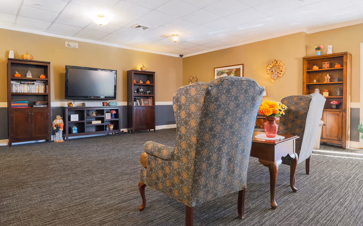 A cozy common living room with upholstered armchairs, a TV on a media console, bookshelves, and a side table with flowers.