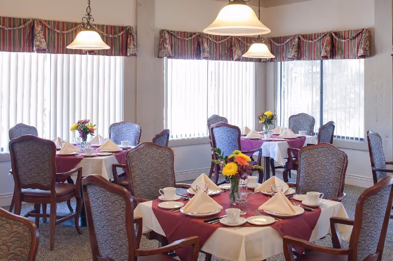 A bright dining room with several round tables covered with white and burgundy tablecloths. Each table is set with white plates, cups, silverware, and folded beige napkins. There are floral centerpieces with yellow and red flowers on each table. The room has large windows with vertical blinds and striped valances. Elegant upholstered chairs surround the tables, and pendant lights hang from the ceiling.