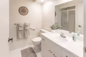 A clean and bright bathroom featuring a white vanity with a sink, a large mirror, a toilet, a glass-enclosed shower, and two striped towels hanging on a towel rack. There is a decorative circular wall piece above the towels and a gray bath mat on the floor.