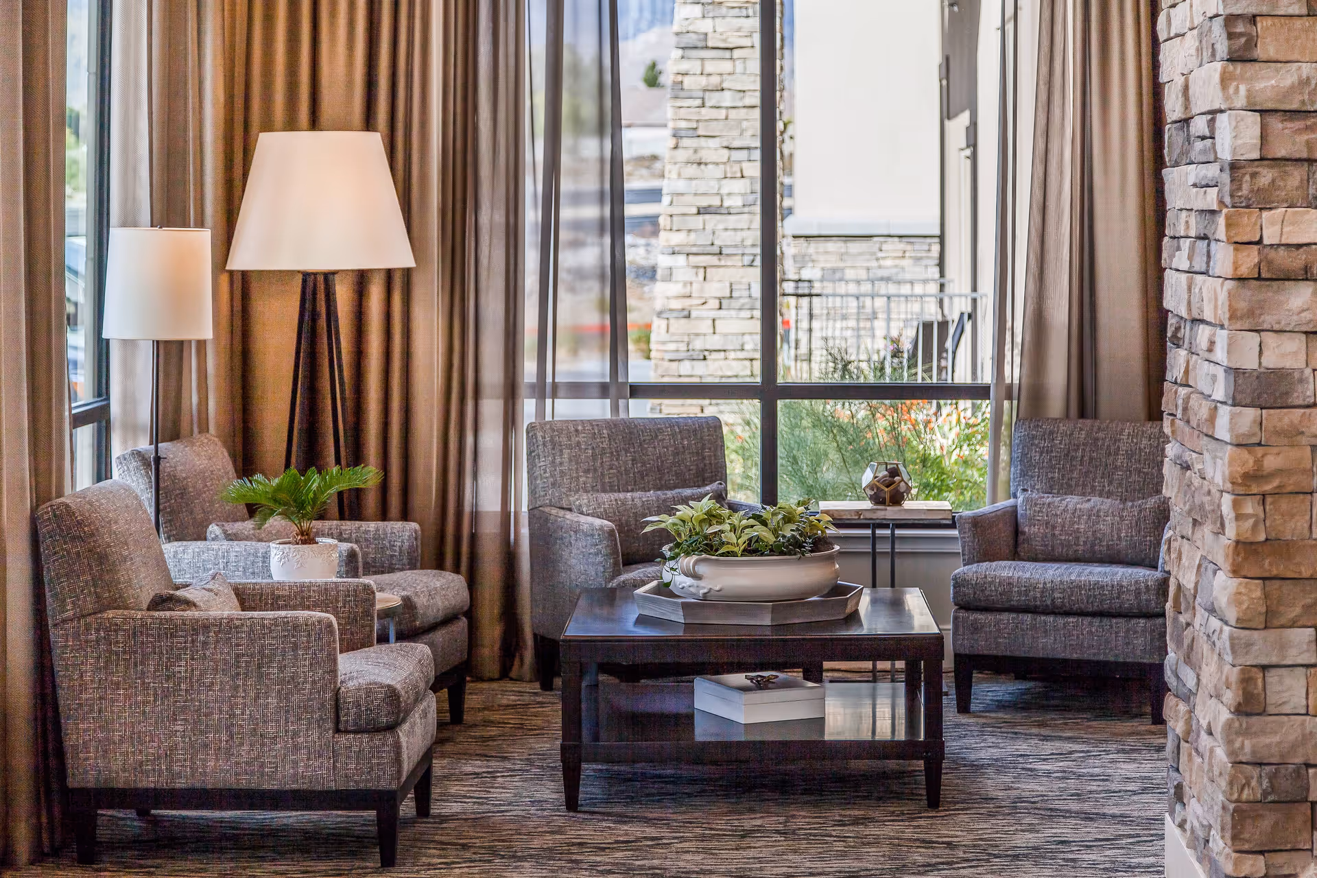 A cozy sitting area with four gray upholstered armchairs arranged around a dark wooden coffee table with a decorative plant centerpiece. Two floor lamps with white shades stand behind the chairs, and large windows with sheer and brown curtains let in natural light. A stone pillar is visible on the right side of the image.