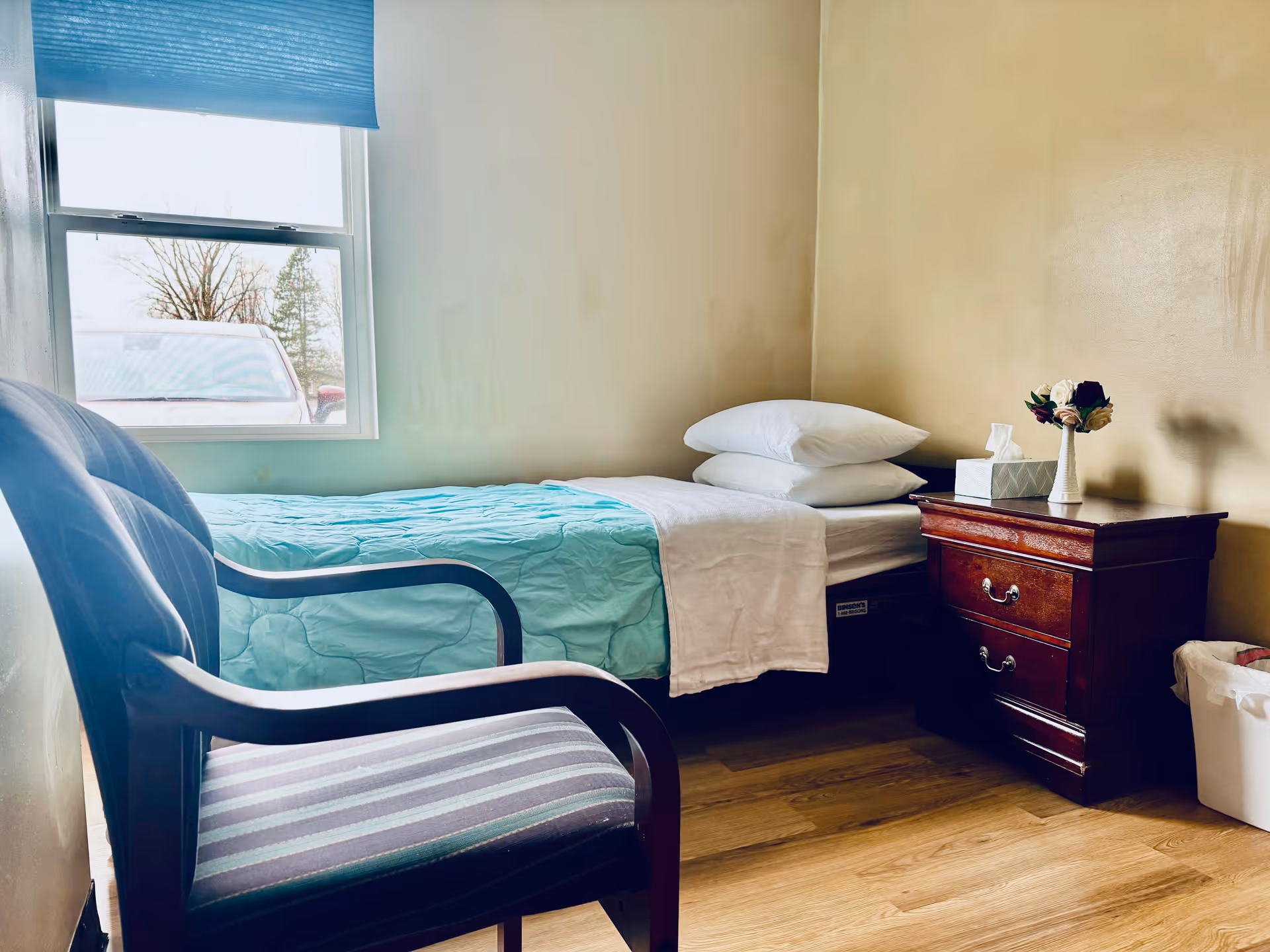 A small bedroom with a single bed made with a light blue quilt and white sheets and pillows. Next to the bed is a wooden nightstand with a tissue box and a small vase with artificial flowers. A striped cushioned chair is placed near the bed. There is a window with a blue blind partially open, showing a parked car and some trees outside. The floor is wooden and the walls are painted beige.