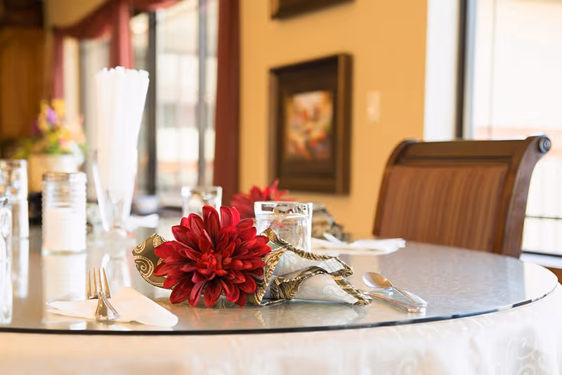 A close-up view of a round dining table set with a glass top, featuring a red flower decoration on a folded napkin, silverware, and glasses. The background shows a softly lit room with framed artwork on the wall and a wooden chair.