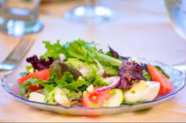 A close-up view of a fresh salad on a clear glass plate, featuring mixed greens, tomato wedges, cucumber slices, red onion, and a boiled egg, placed on a table with a fork and glass of water in the background.
