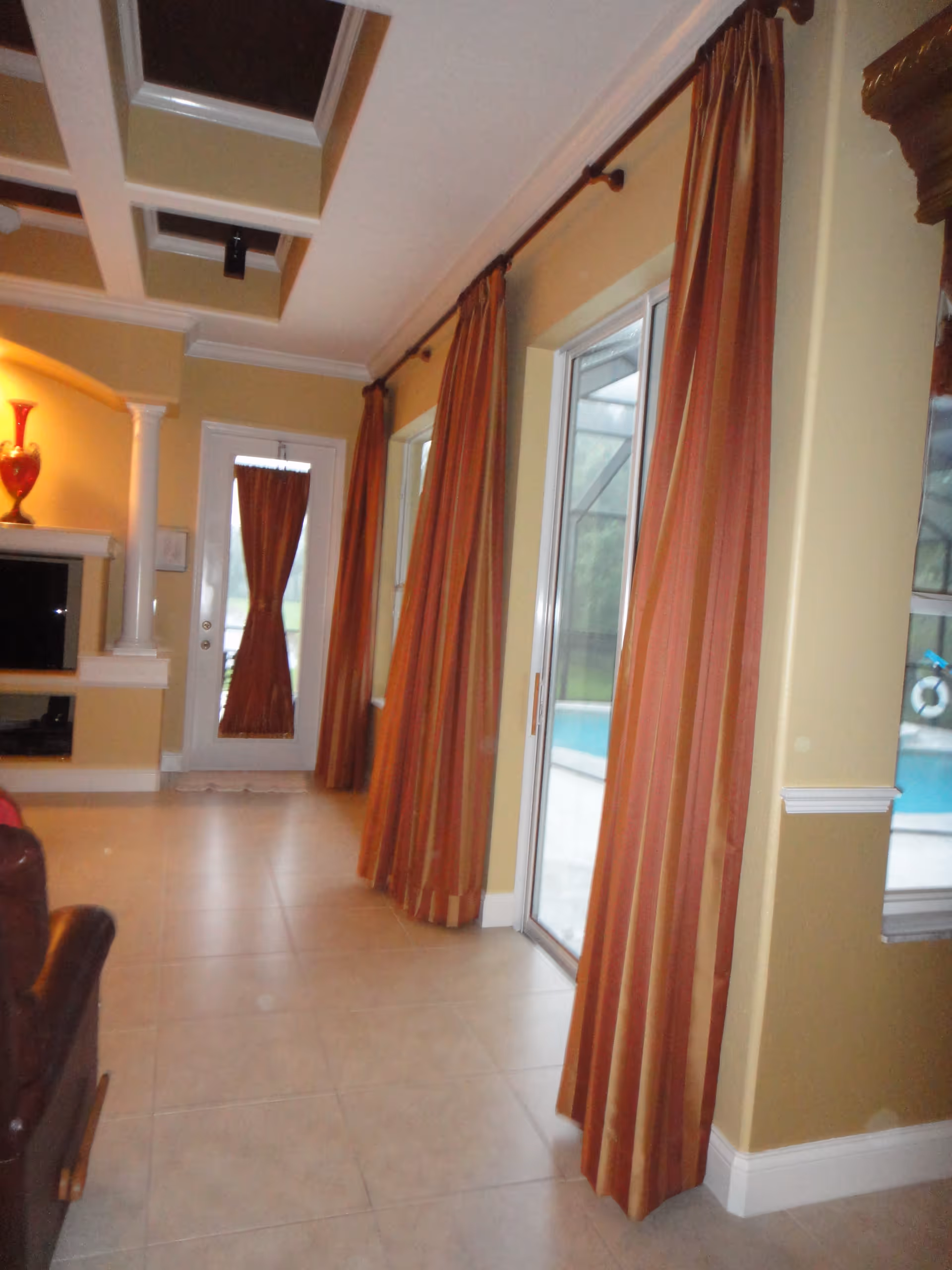 Interior view of a living room area with beige tiled floor, yellow walls, and large windows covered with long orange and red striped curtains. There is a glass sliding door leading outside to a pool area. A white door with a small curtain is visible at the end of the room. The ceiling has a recessed design with white beams. A red decorative vase is placed on a shelf near a TV.