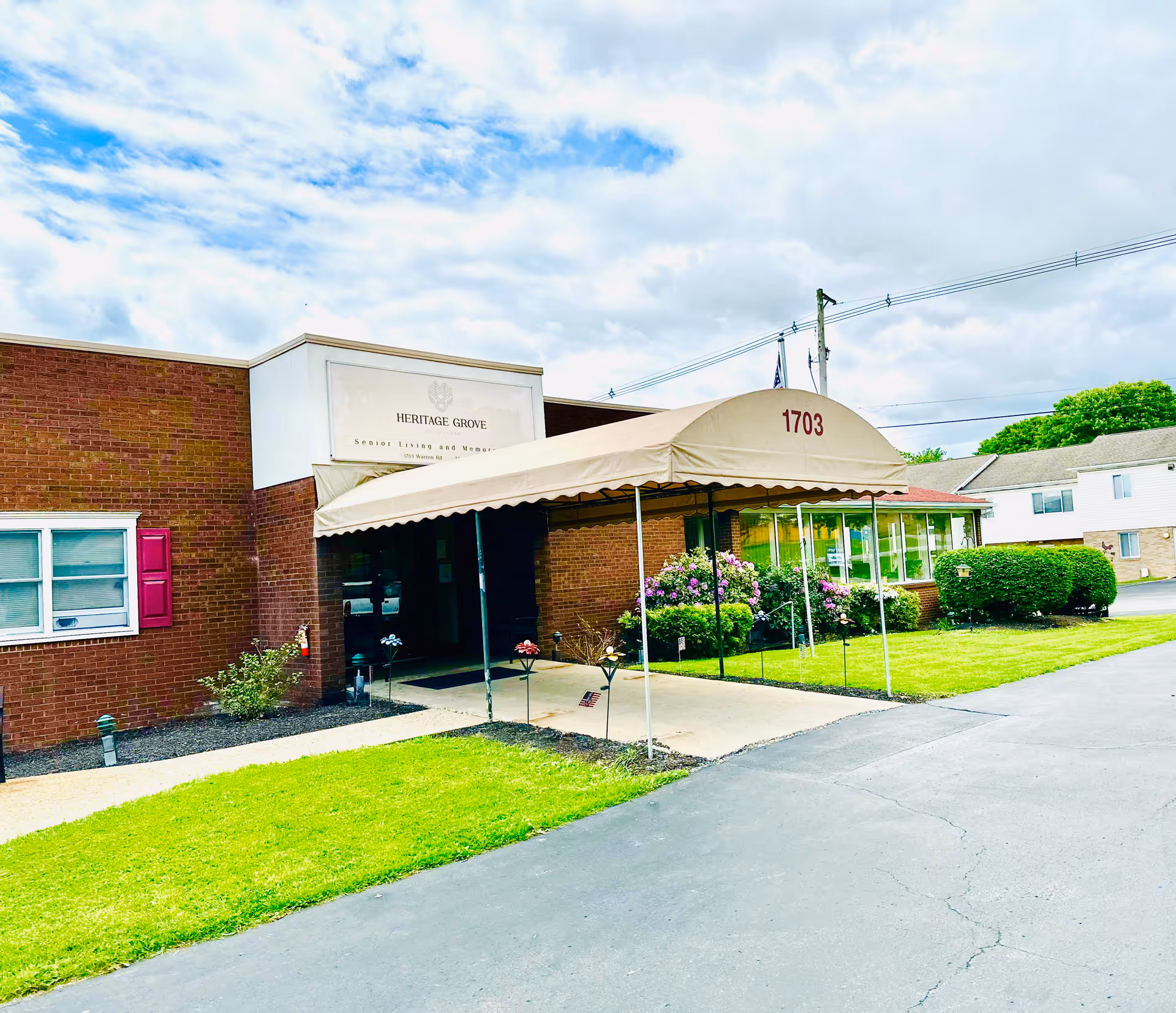 Exterior view of Heritage Grove at Indiana, a senior living and memory care facility. The building is made of red brick with a beige canopy over the entrance displaying the number 1703. There are green lawns, bushes, and flowers around the entrance, and a partly cloudy sky overhead.